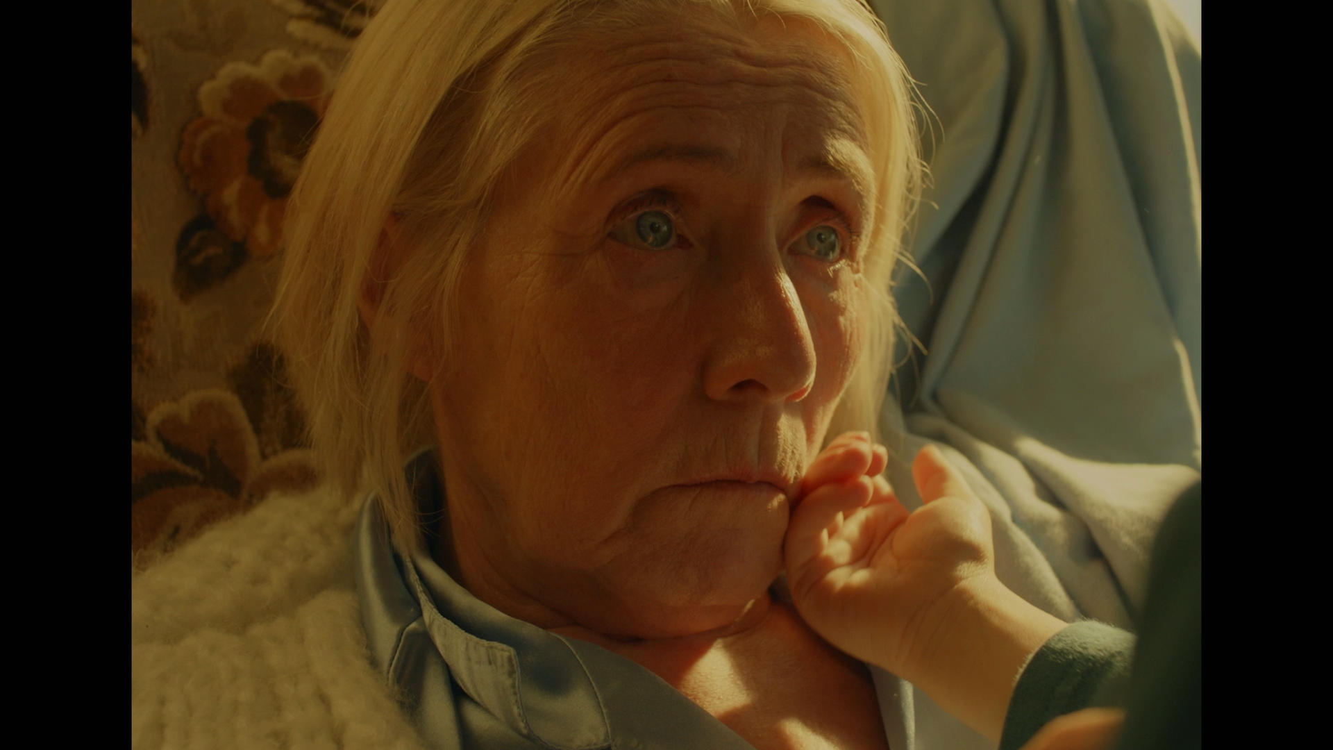 Close-up of an elderly woman with blonde hair, lying in bed, looking contemplative with a hand lightly touching her chin.
