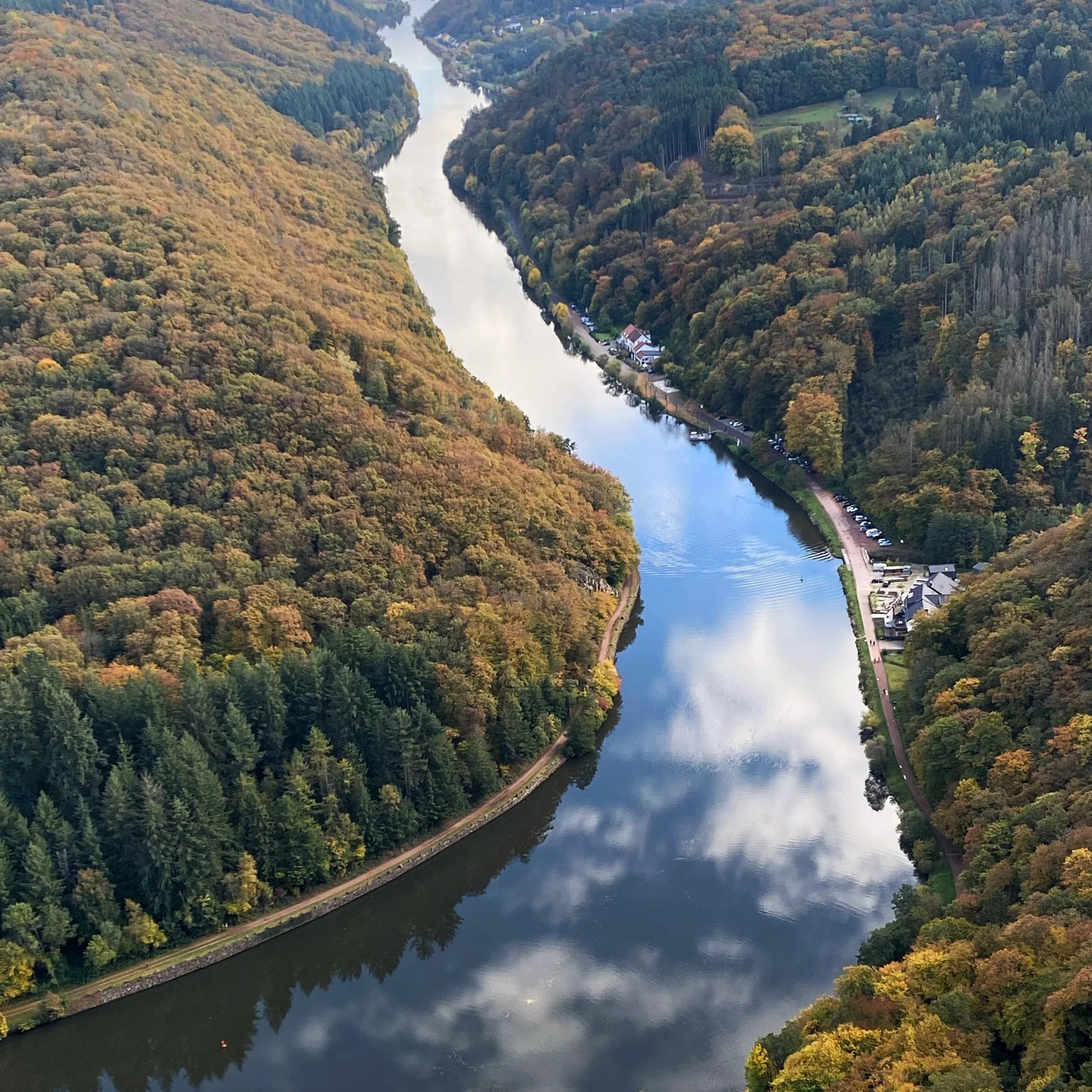 Une vue aérienne d'une rivière sinuante entourée de collines et de forêts aux feuilles d'automne, avec quelques bâtiments et une route au bord de l'eau.