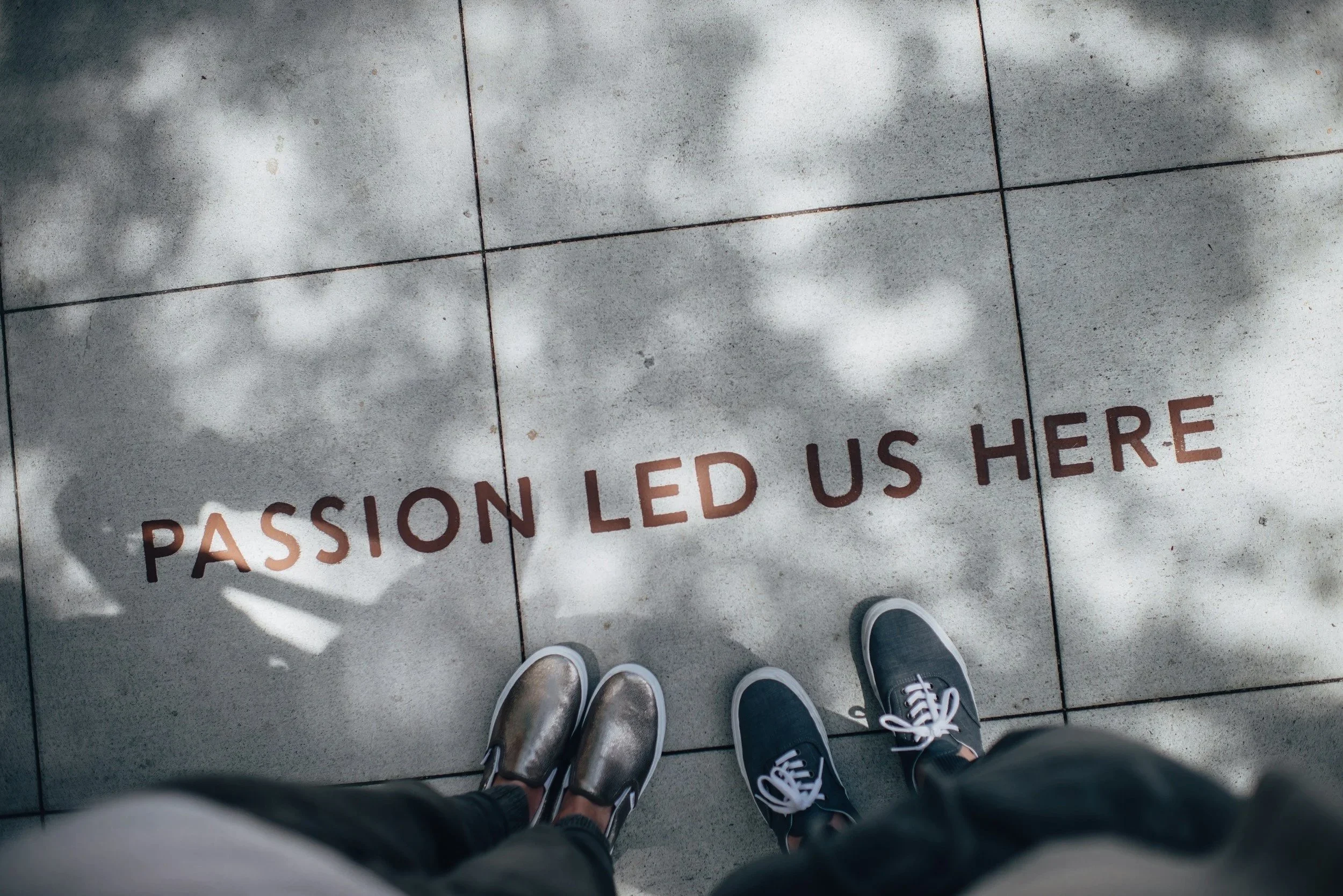 Shoes worn by the person standing on a tiled floor with shadows of trees and the words 'Passion Led Us Here' written on the tiles.