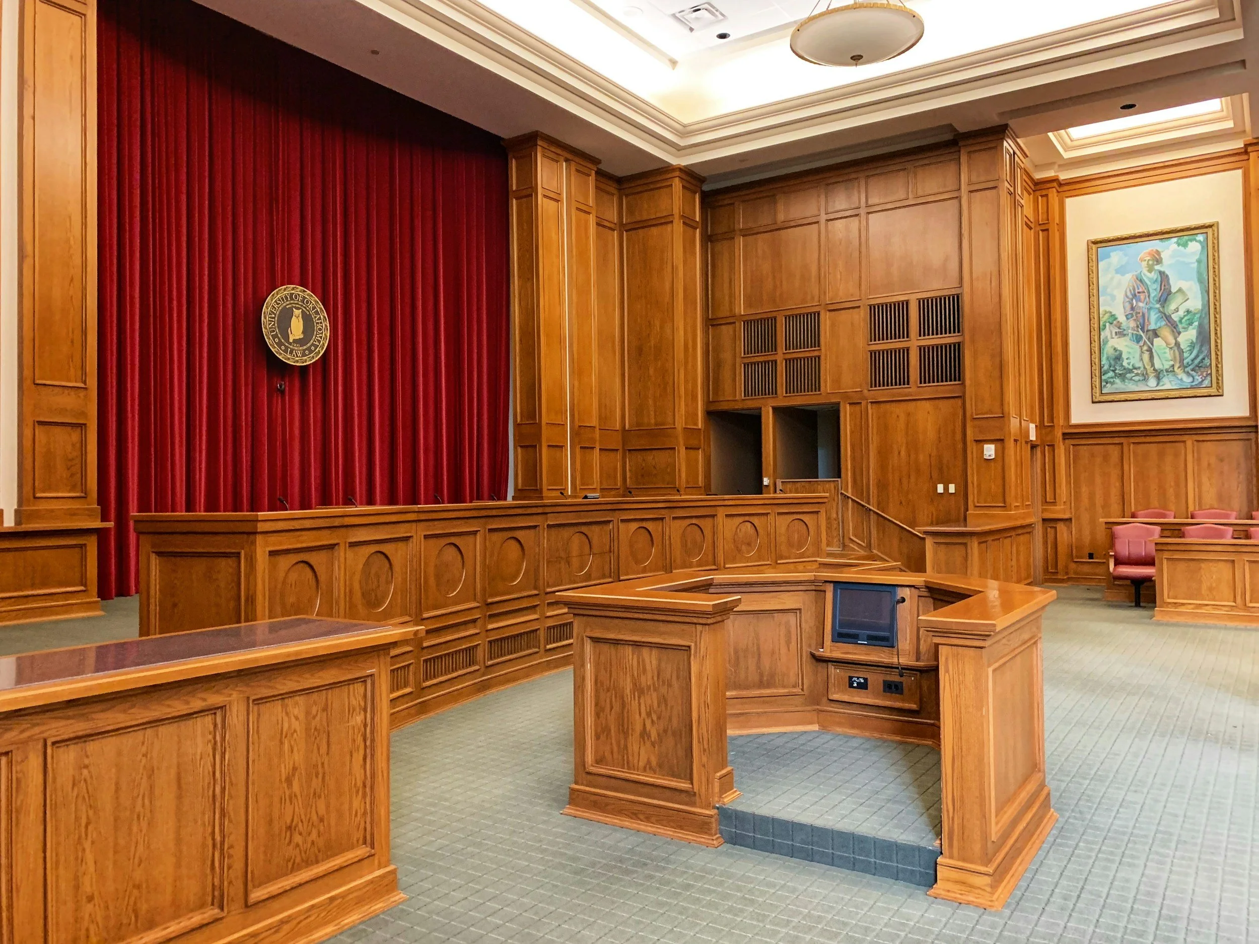 Empty courtroom with wooden paneling, a red curtain with an emblem, and a portrait of a person in regal attire.
