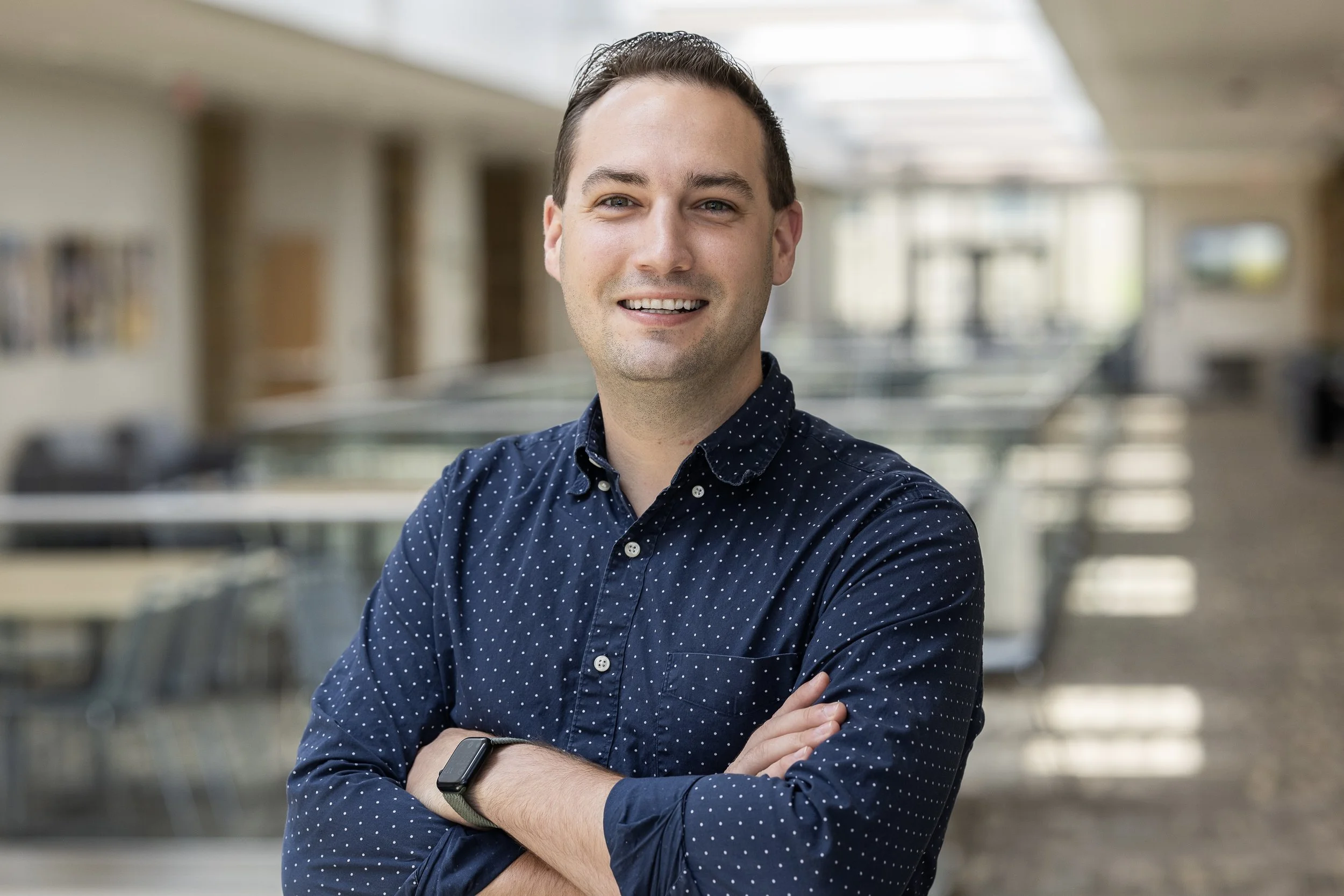 A smiling man with dark hair, wearing a navy blue polka dot shirt and a smartwatch, standing with arms crossed in a well-lit indoor space with blurred background.