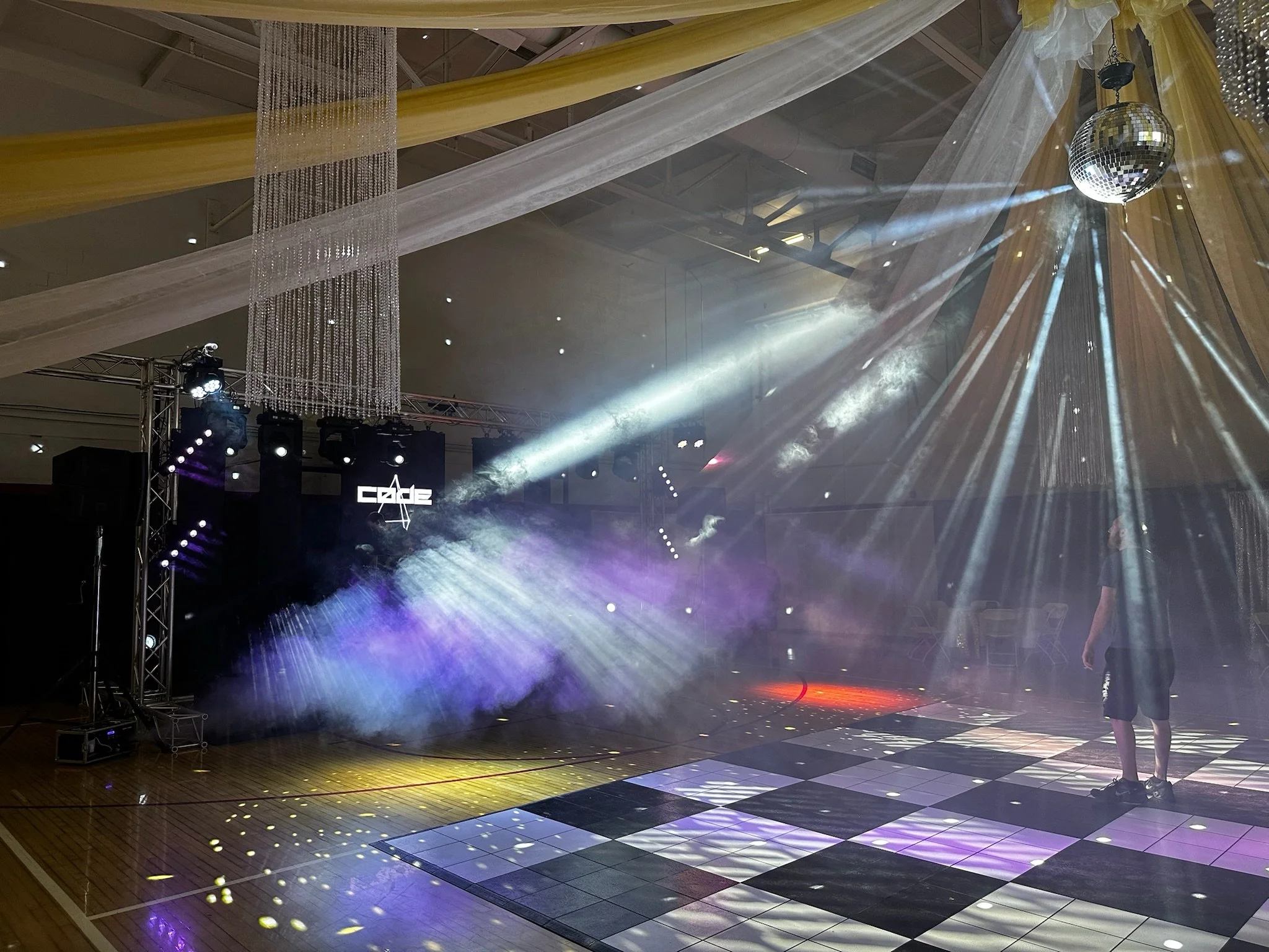 An empty dance floor with purple and yellow stage lights and a disco ball hanging from the ceiling. There is fog and colorful lighting effects on the floor, with a person standing near the right side of the dance floor.