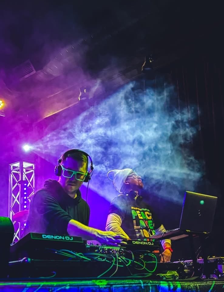 Two DJs are performing on stage with colorful lights and smoke effects. One DJ is wearing neon glasses and a hoodie, while the other is in a beanie and glasses with a T-shirt that says 'PEACE LOVE HAPPINESS'. Both are using DJ equipment and appear to be mixing music.