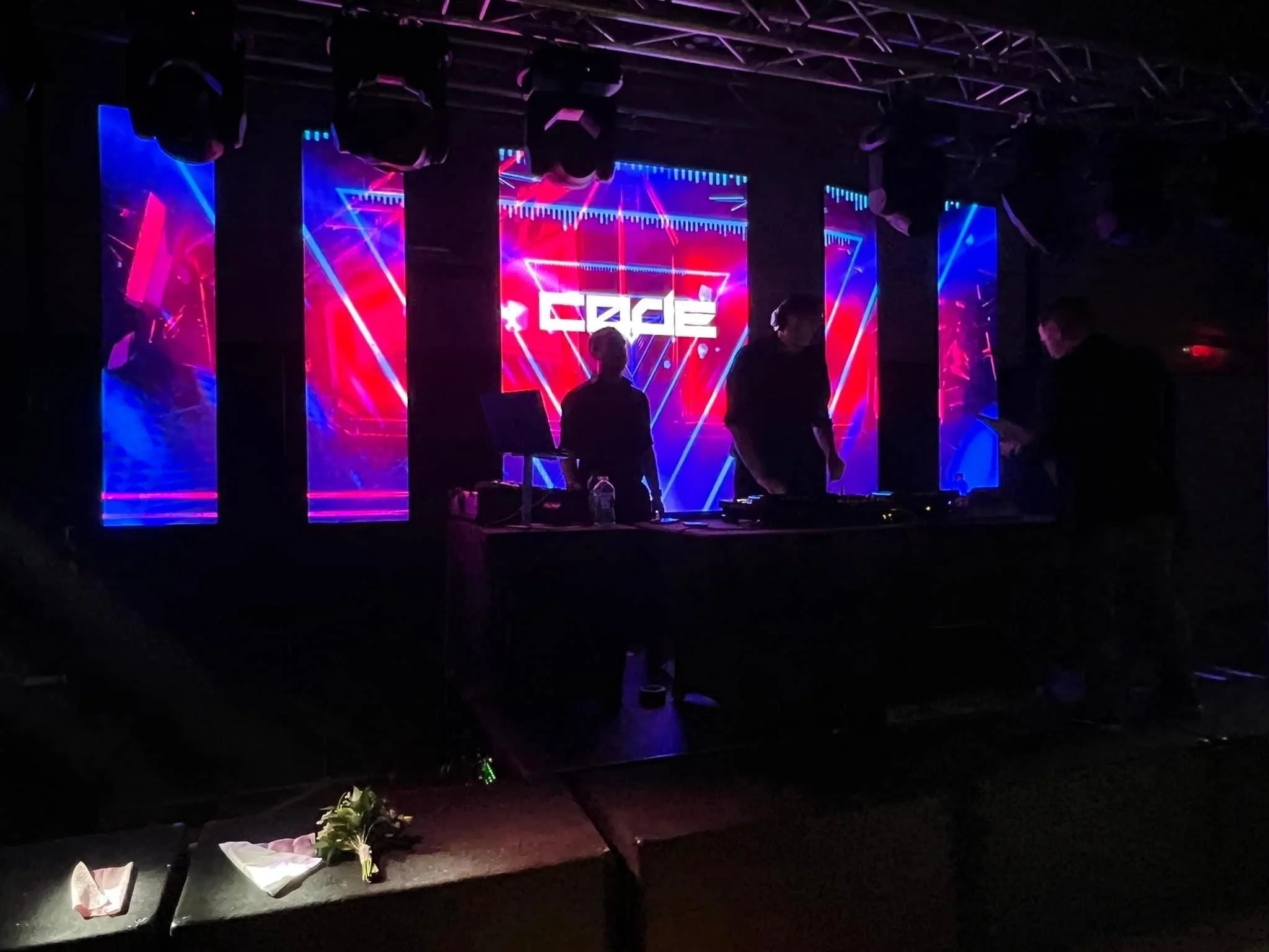 A DJ performing on stage with a vibrant digital background displaying abstract graphics and the word 'CACE.' Three people are working at the DJ booth, with one in the center and two on either side. The stage is lit with purple and blue lights, and there are large hanging screens behind them showing matching visuals.