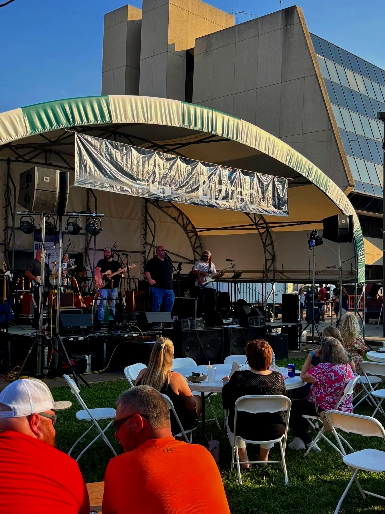 Live outdoor concert featuring a band performing on a stage with a curved canopy. Audience members are seated at tables and chairs on the grass, watching the performance, with a modern building in the background.