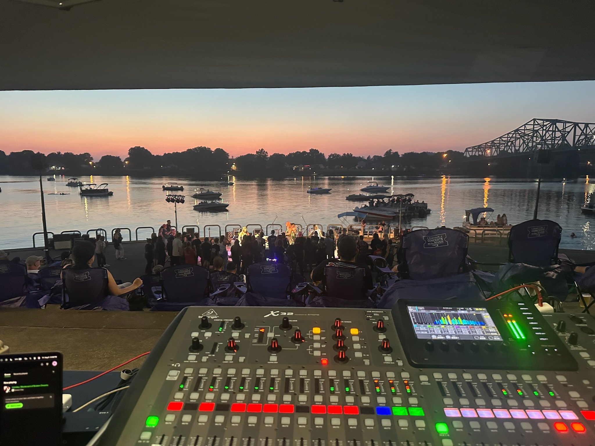 View of a river at sunset with boats anchored, people gathered near the shore, and a bridge in the background. The image is taken from a stage with a music mixer in the foreground.