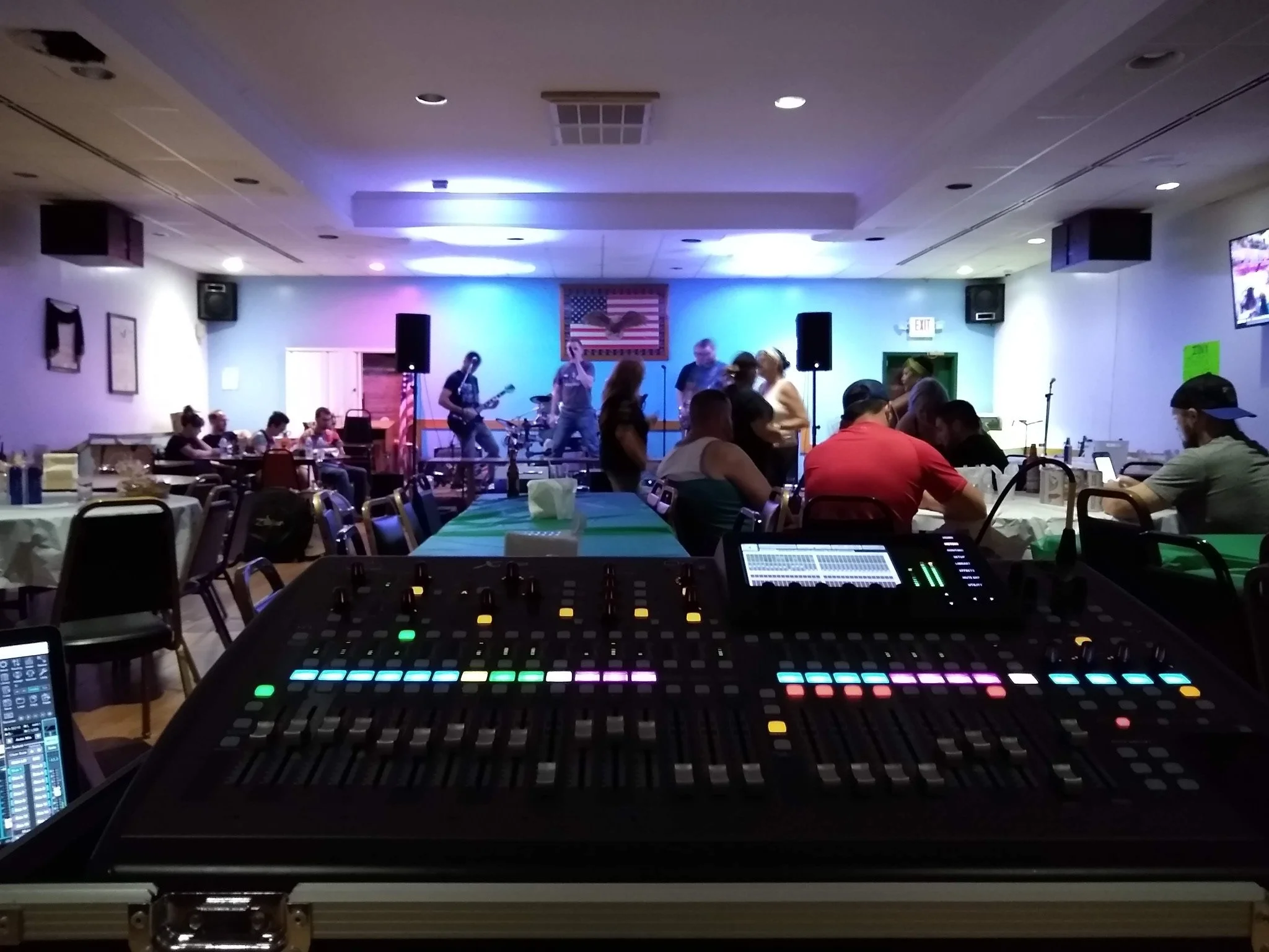 View from behind a sound mixing board in a large event room with people seated at tables and a band performing on stage with blue and purple lighting, American flag, and wall-mounted TV screens.
