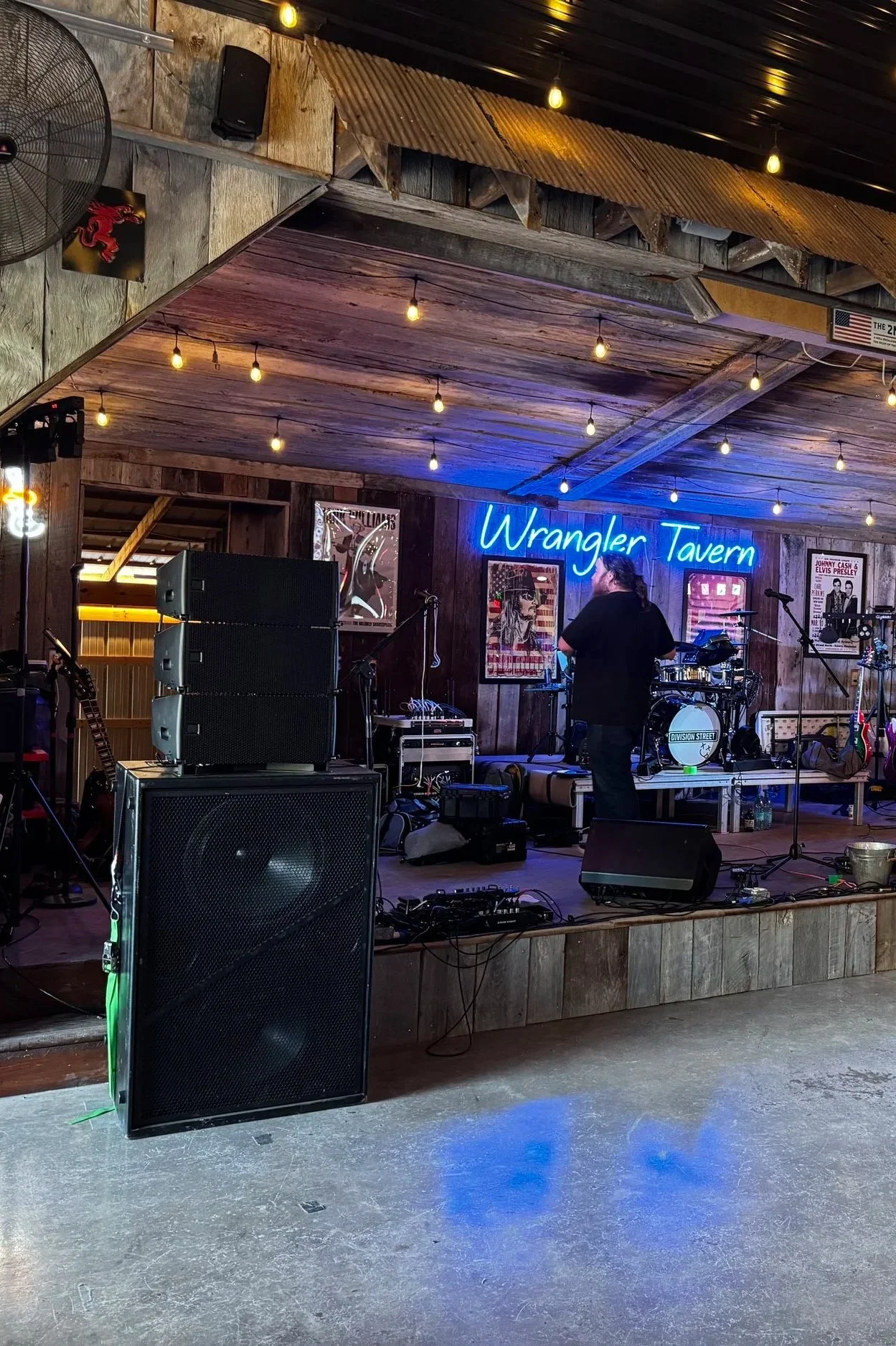 Stage at Wrangler Tavern with musical instruments, line array JBL SRX speakers, and sound equipment, decorated with stage lights and posters on wooden walls, preparing for a performance. Sound Company.