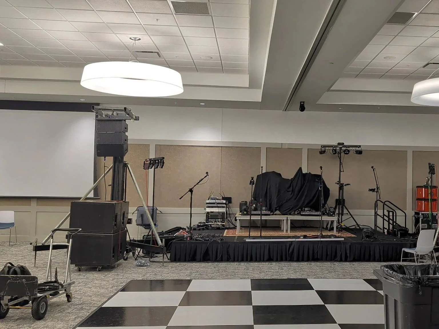 Empty event stage with microphones, line array JBL SRX speakers, and Chauvet lighting equipment, covered with black cloth, set in a large conference room with a black and white checkered dance floor. Sound Company.