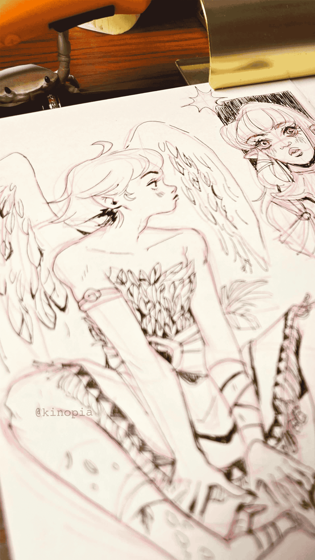 Sketch of a fantasy character with wings, long hair, and patterned clothing, drawn on paper with a pen, on a wooden desk.