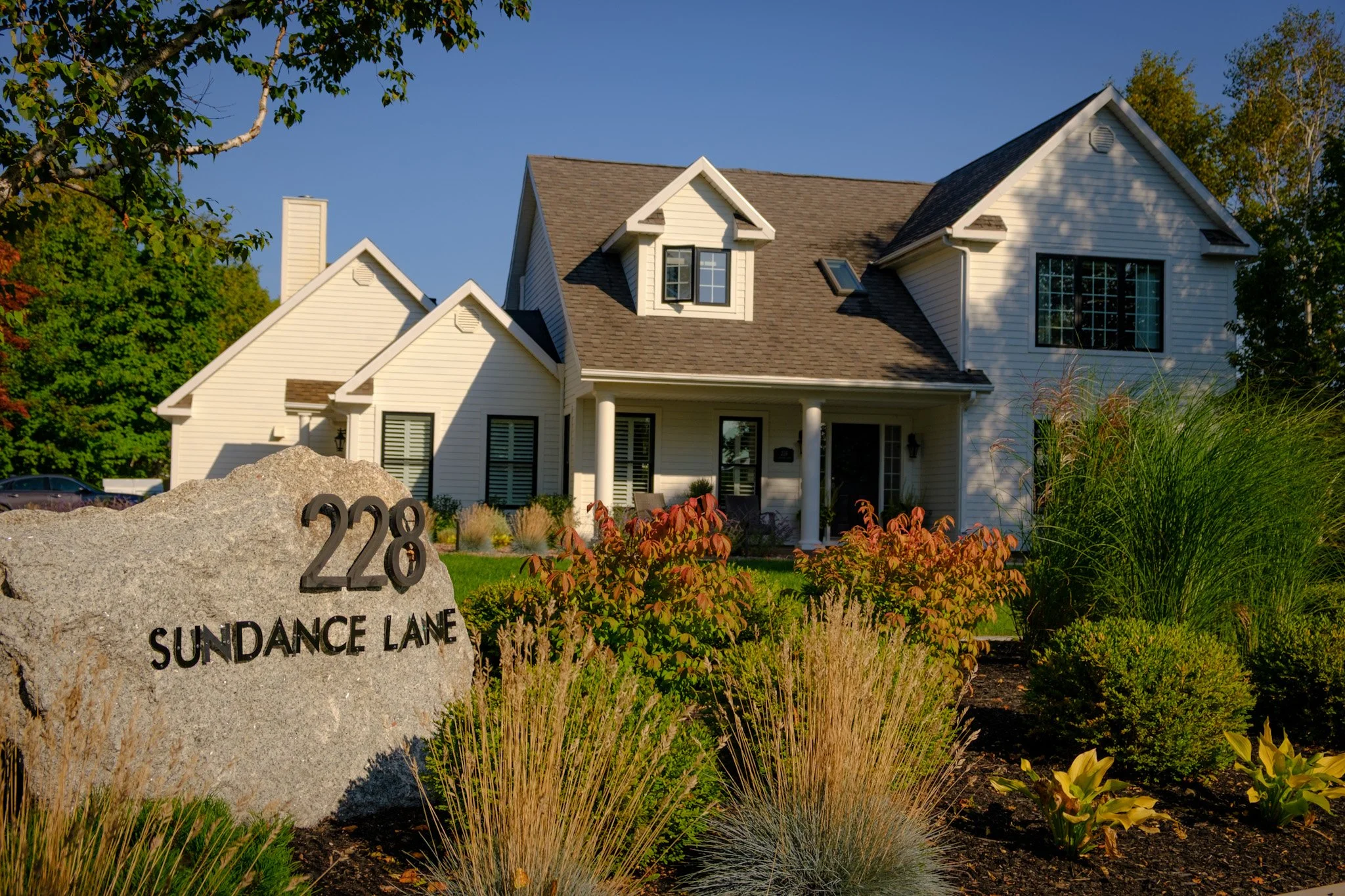 A white two-story house with a porch, surrounded by well-kept garden and bushes, with the address 228 Sundance Lane on a large stone marker in the foreground, under a clear blue sky.