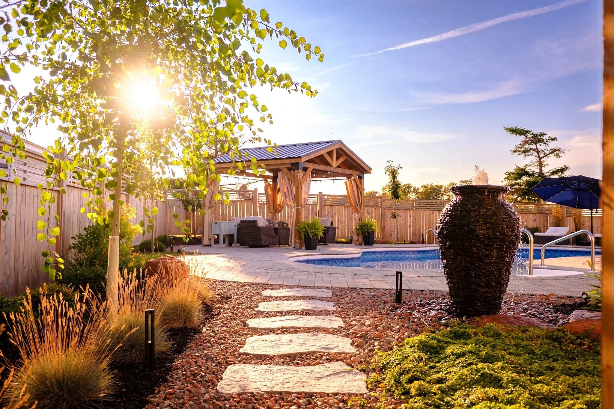 A backyard with a swimming pool, a wooden gazebo with curtains, outdoor furniture, a large decorative fountain, and a paved stone pathway surrounded by landscaped plants and trees, illuminated by warm sunlight.