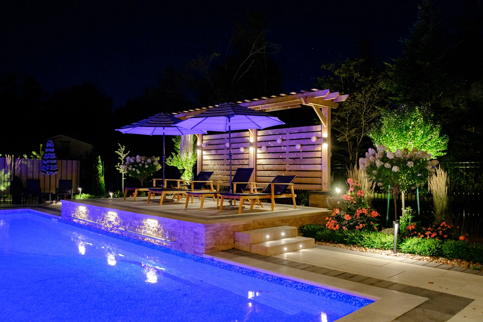 Night view of a backyard with a swimming pool, illuminated by underwater lights, and a wooden poolside deck with lounge chairs and umbrellas, surrounded by landscaped plants and flowers.