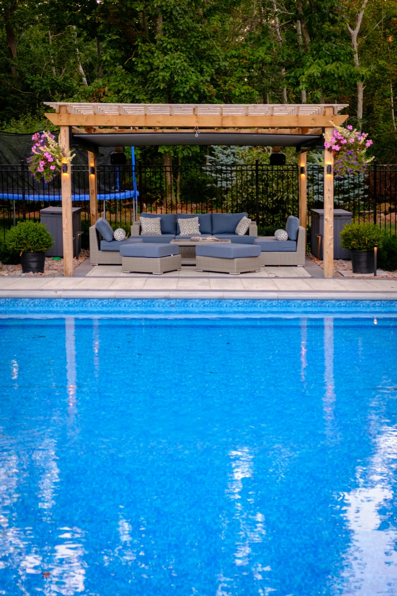A backyard with a swimming pool, a wooden pergola with outdoor seating, potted plants, hanging flower baskets, and a forested background.