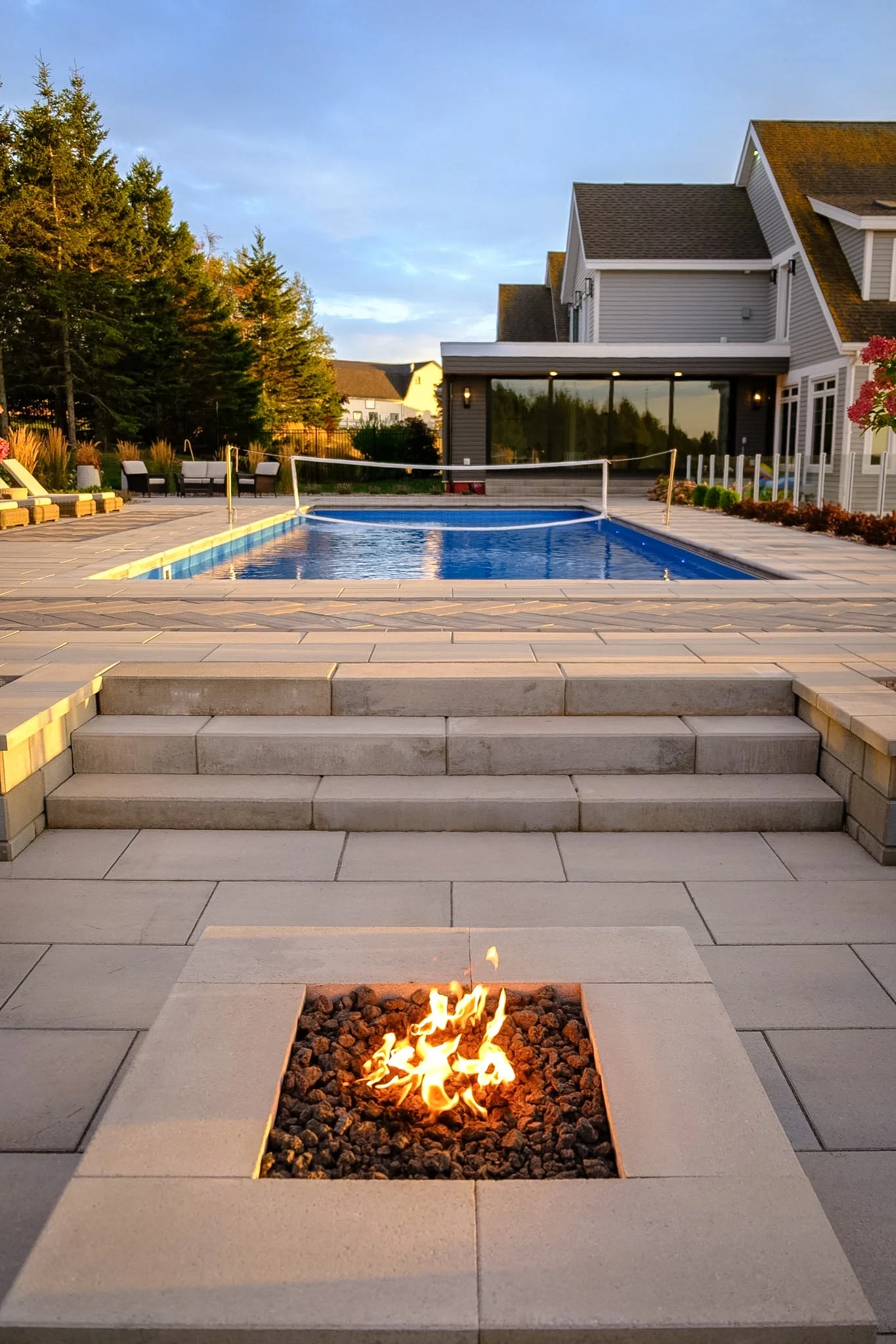 Backyard patio with a small fire feature, steps leading to a swimming pool, surrounded by lounge chairs, patio furniture, and landscaped garden beyond.