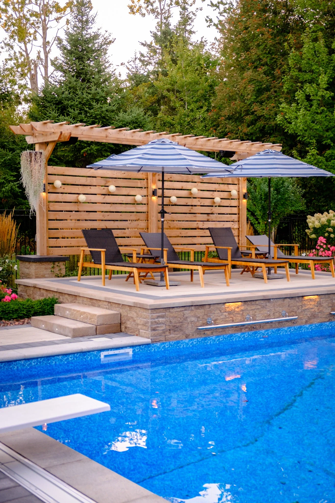 Backyard patio with pool, four black lounge chairs, striped blue umbrellas, string lights, wooden fencing, muita background trees, and a wooden pergola.