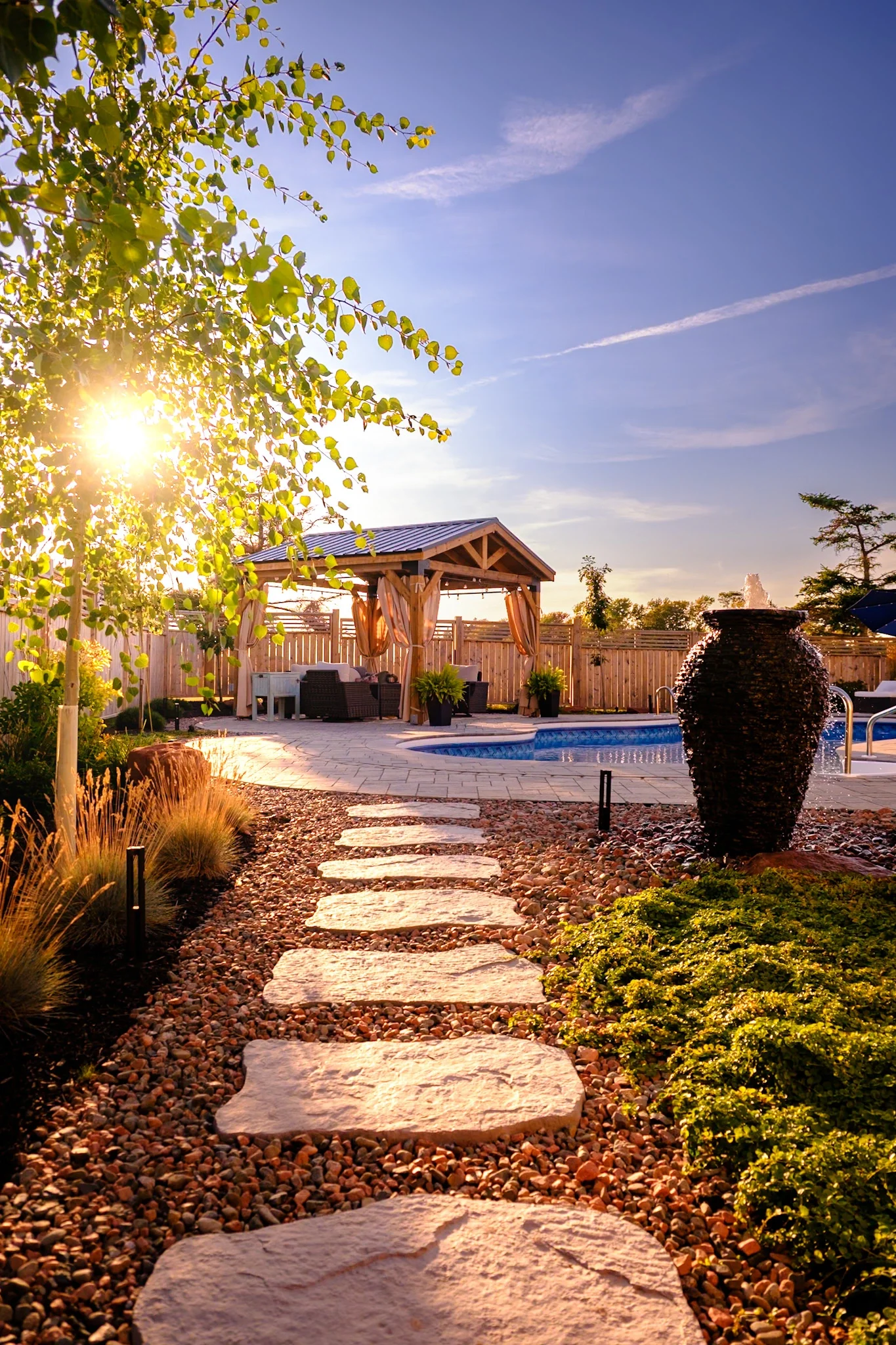 Sunset view of a backyard with a stone pathway leading to a poolside patio area, featuring a wooden gazebo with curtains, outdoor seating, potted plants, and a decorative water fountain, under a clear sky.