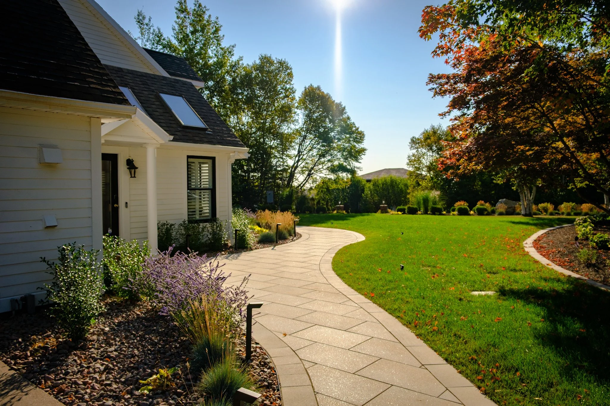 A paved curved walkway runs through a well-maintained lawn with trees and shrubs on a sunny day.