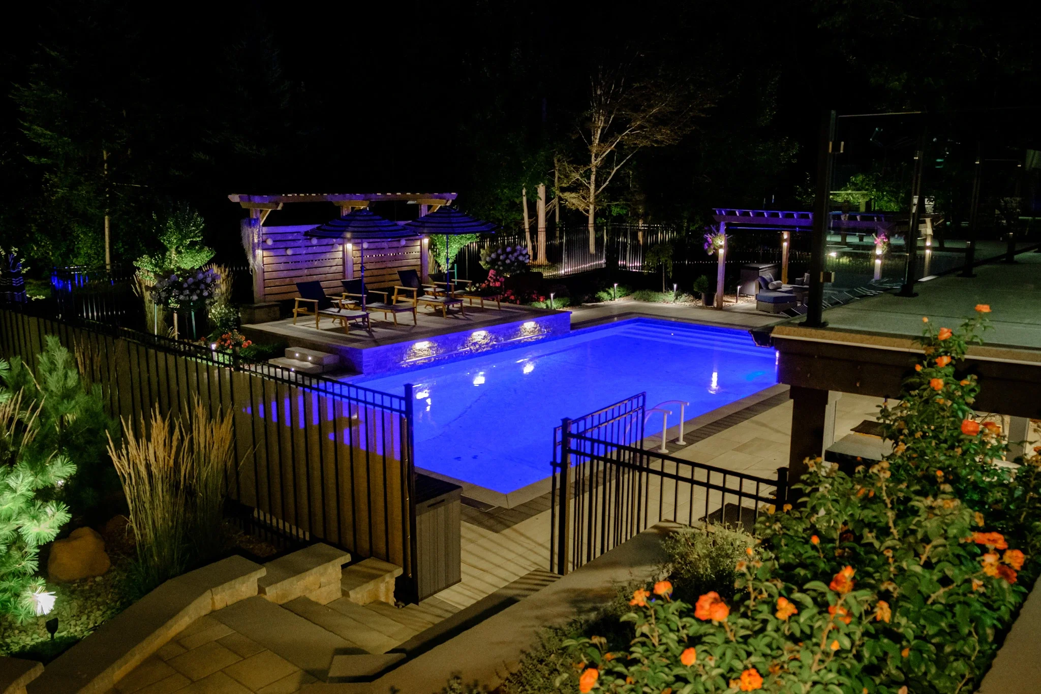 Night view of a backyard with a lit swimming pool surrounded by a fence, with outdoor seating areas, a wooden pergola with chairs and umbrellas, trees, and garden lights.