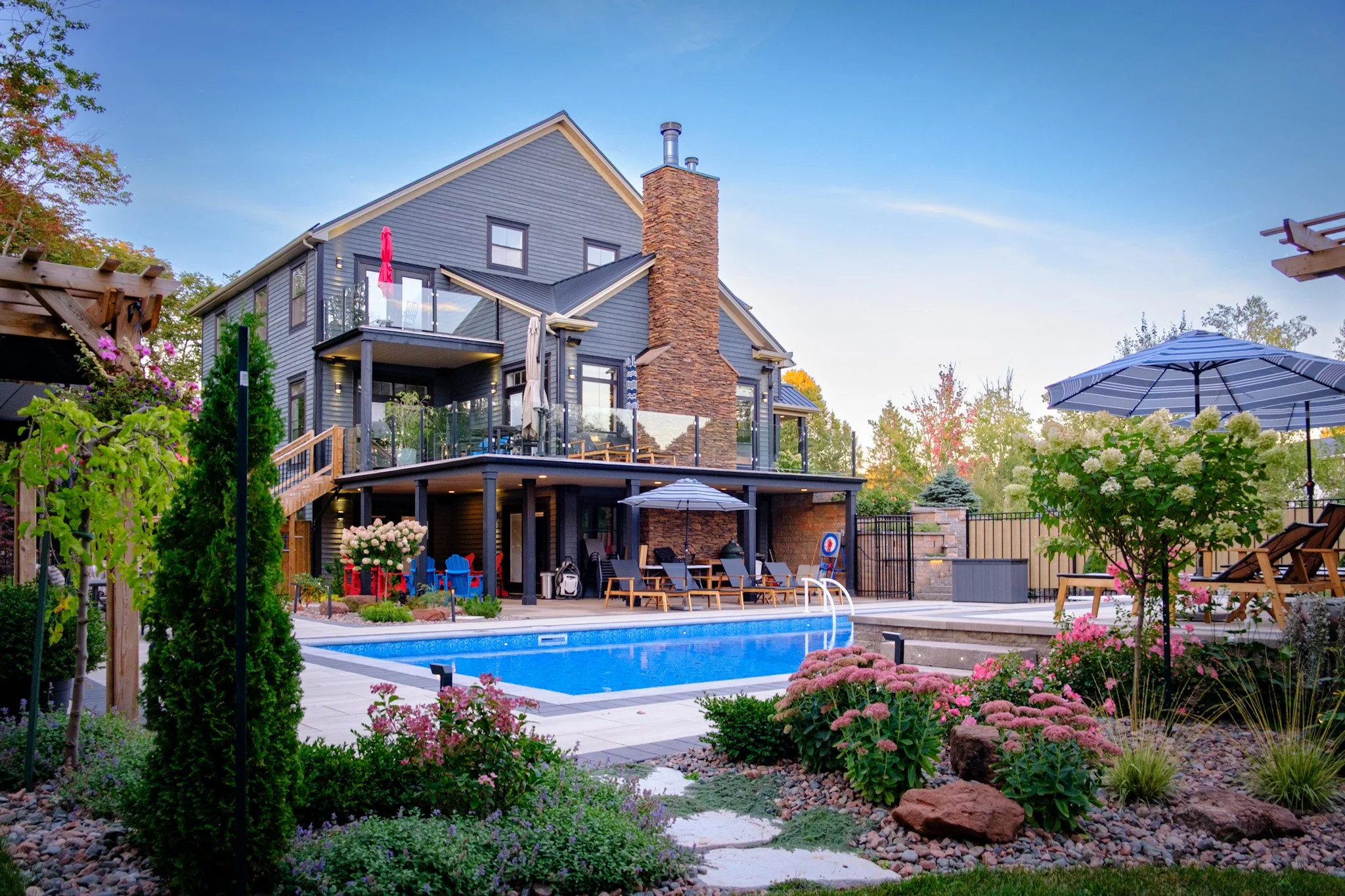 Backyard with swimming pool, lounge chairs, umbrellas, and a two-story house with a stone chimney and balconies, surrounded by colorful plants and flowers.