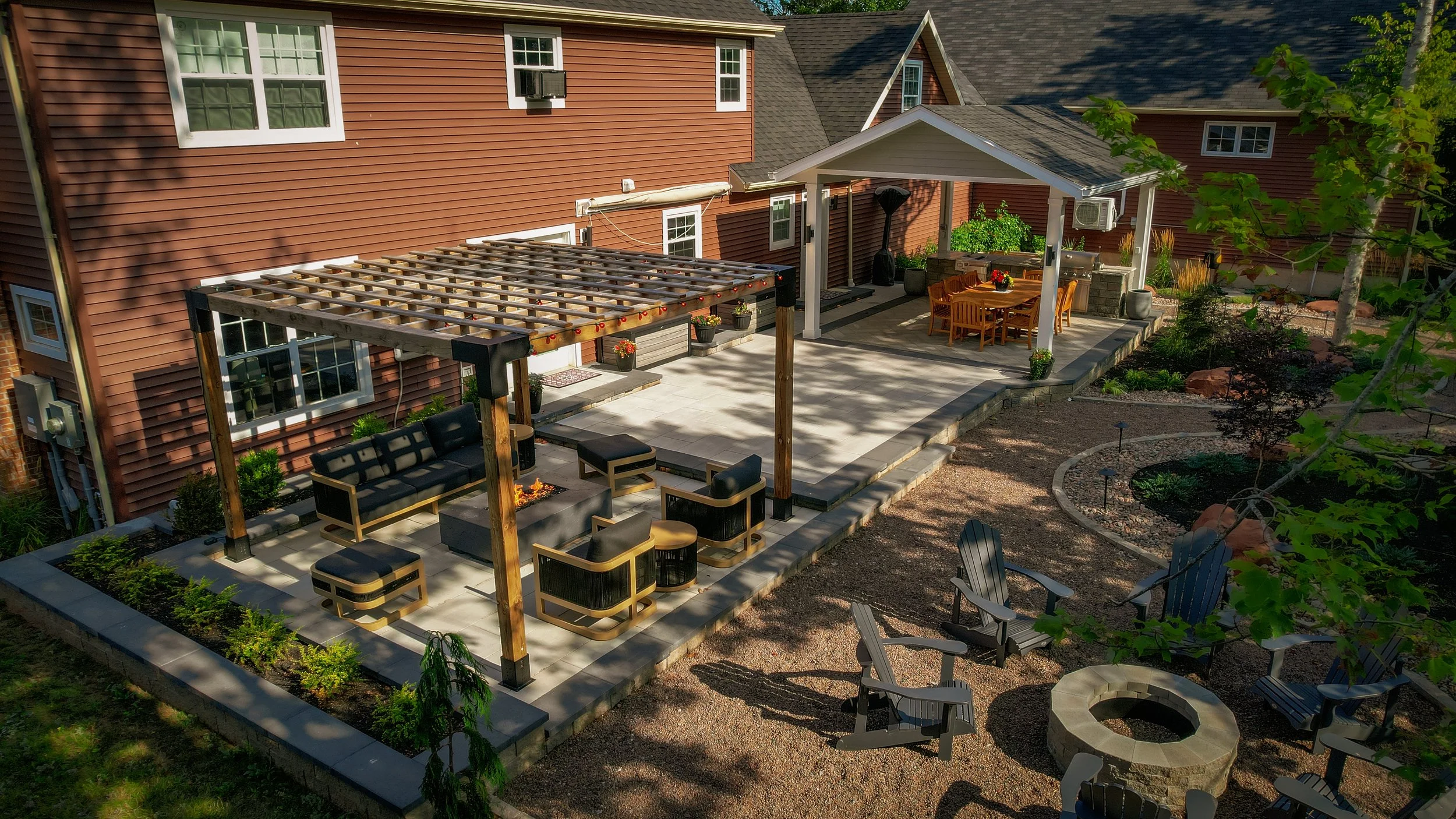 A backyard patio with a pergola, outdoor furniture, and a dining area under a pavilion. There are chairs and a fire pit with chairs around it, and landscaped garden beds.