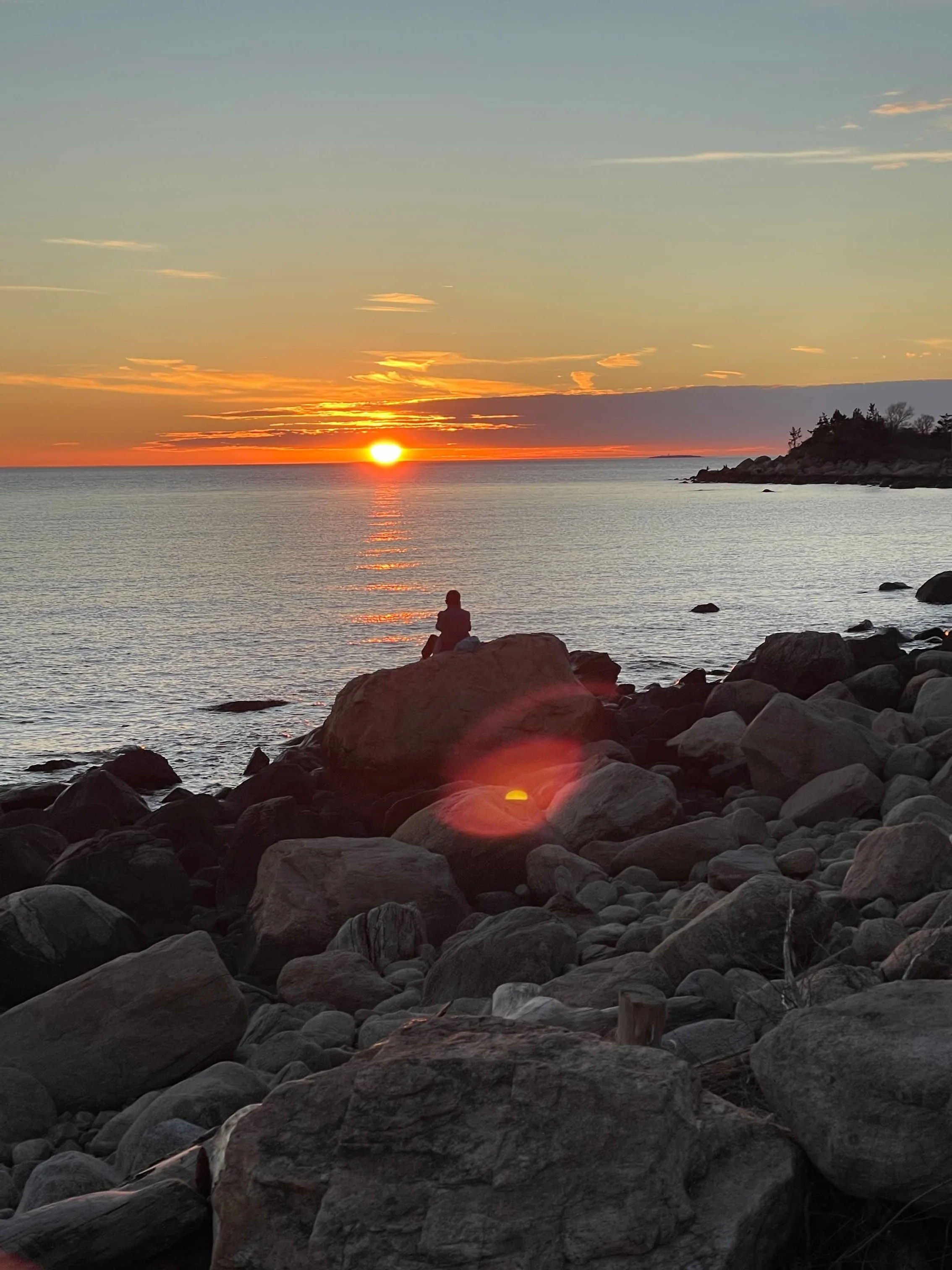Sunset over a rocky beach with calm water and a person sitting on a large rock.