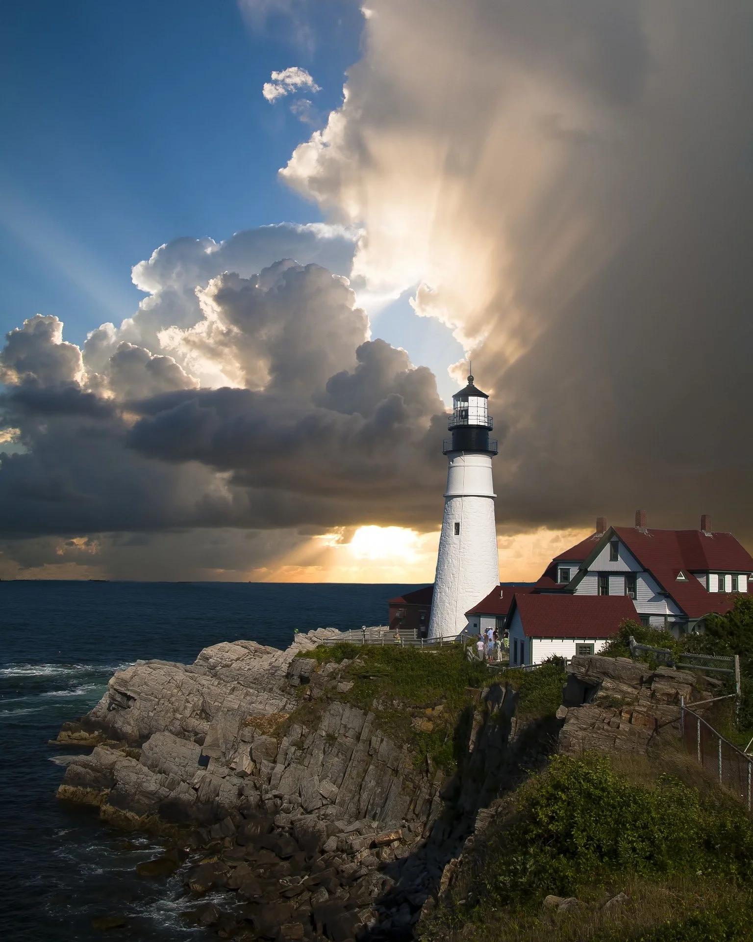 A lighthouse on rocky coast near the ocean at sunset with dramatic clouds overhead.