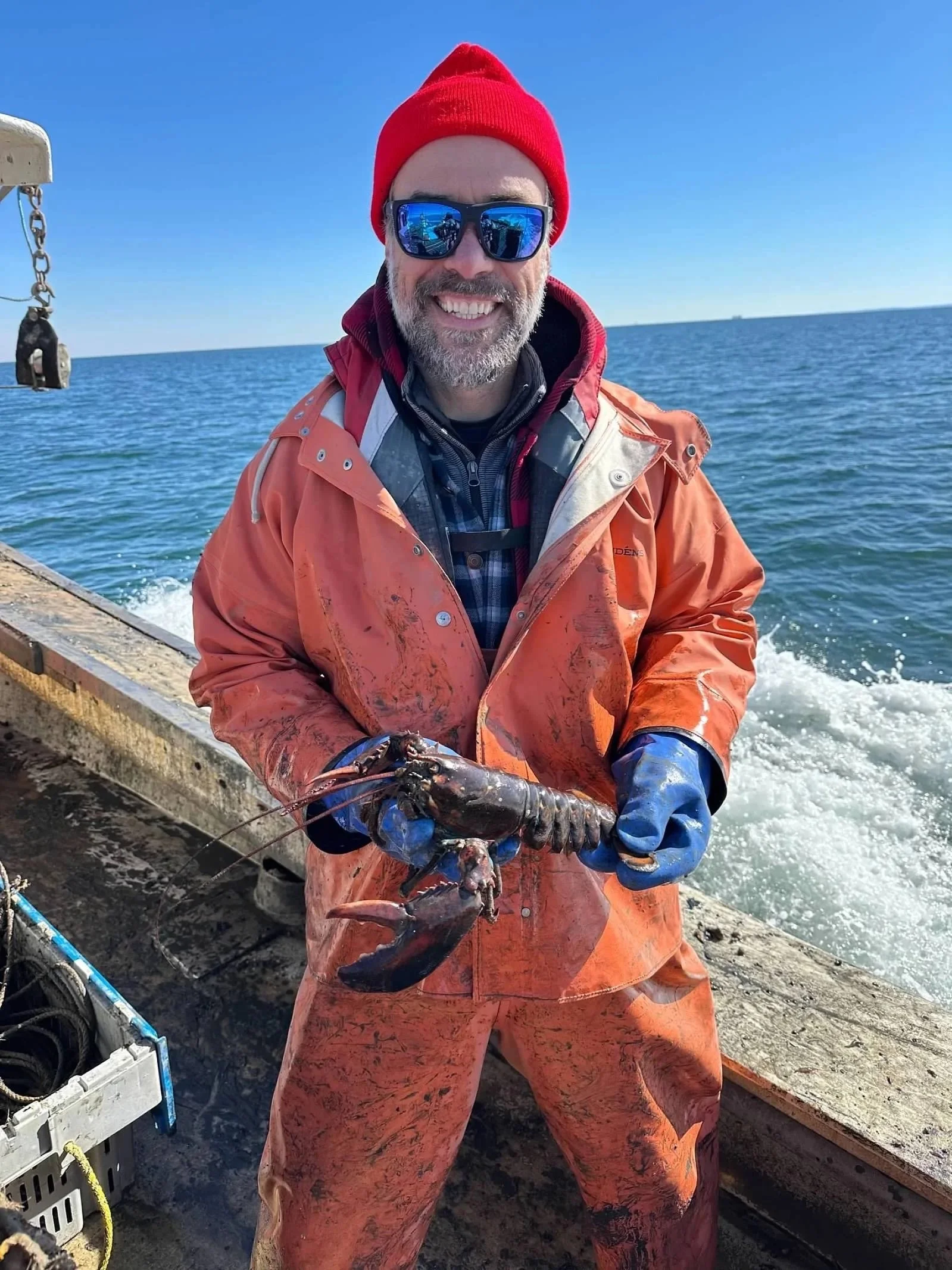 Man in orange waterproof gear, red beanie, and sunglasses holding a lobster on a boat by the ocean with a clear blue sky.