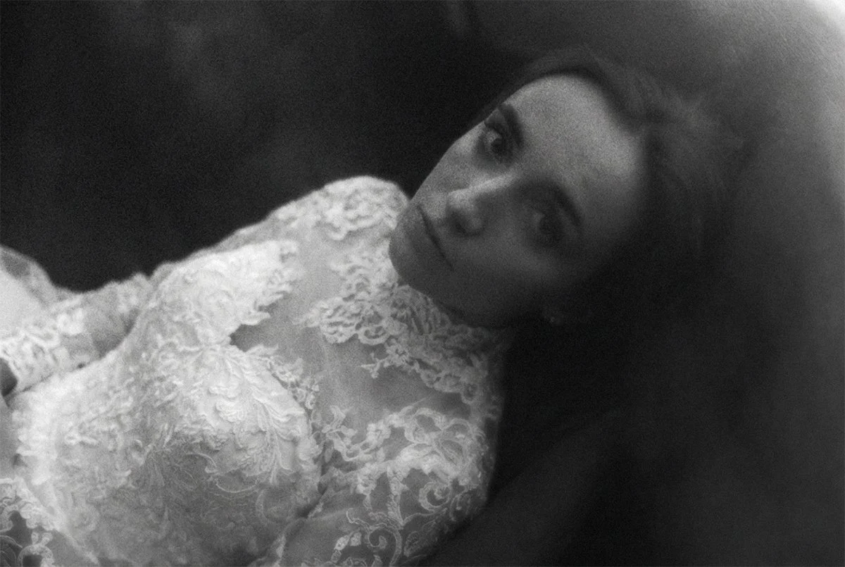 Schwarze und weiße Fotografie eines Mädchens mit langen Haaren, das in einem weißen, Spitzenkleid liegt und direkt in die Kamera schaut.