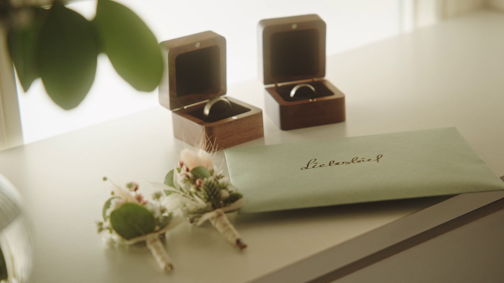 Ein Briefumschlag mit der Aufschrift »Liebesbrief« liegt auf einem weißen Tisch, daneben befinden sich zwei kleine Blumenarrangements. Im Hintergrund sind zwei offene Ringdosen, in denen Eheringe aufbewahrt werden.