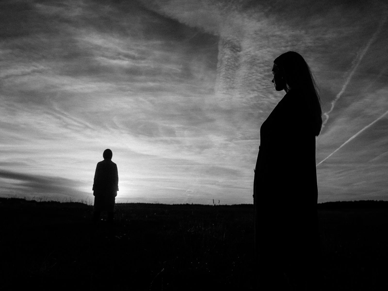 zwei silhouettierte Personen, eine weiblich, die andere männlich, stehen in einer offenen Landschaft bei Sonnenuntergang, Himmel mit Wolken. Ästhetische Hochzeitsfotografie