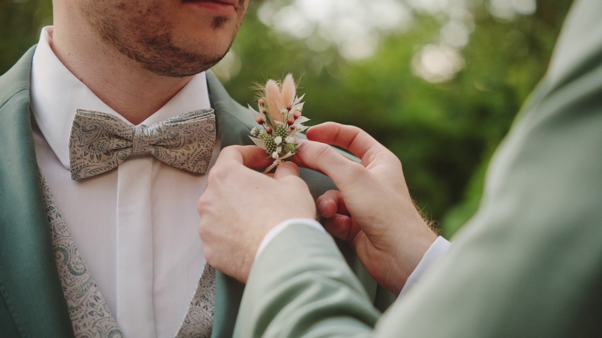 Person steckt einem anderen Person eine Blumenkombination an die Krawatte, bei einer Hochzeit, im Freien mit grüner, unscharfer Hintergrund. Getting ready