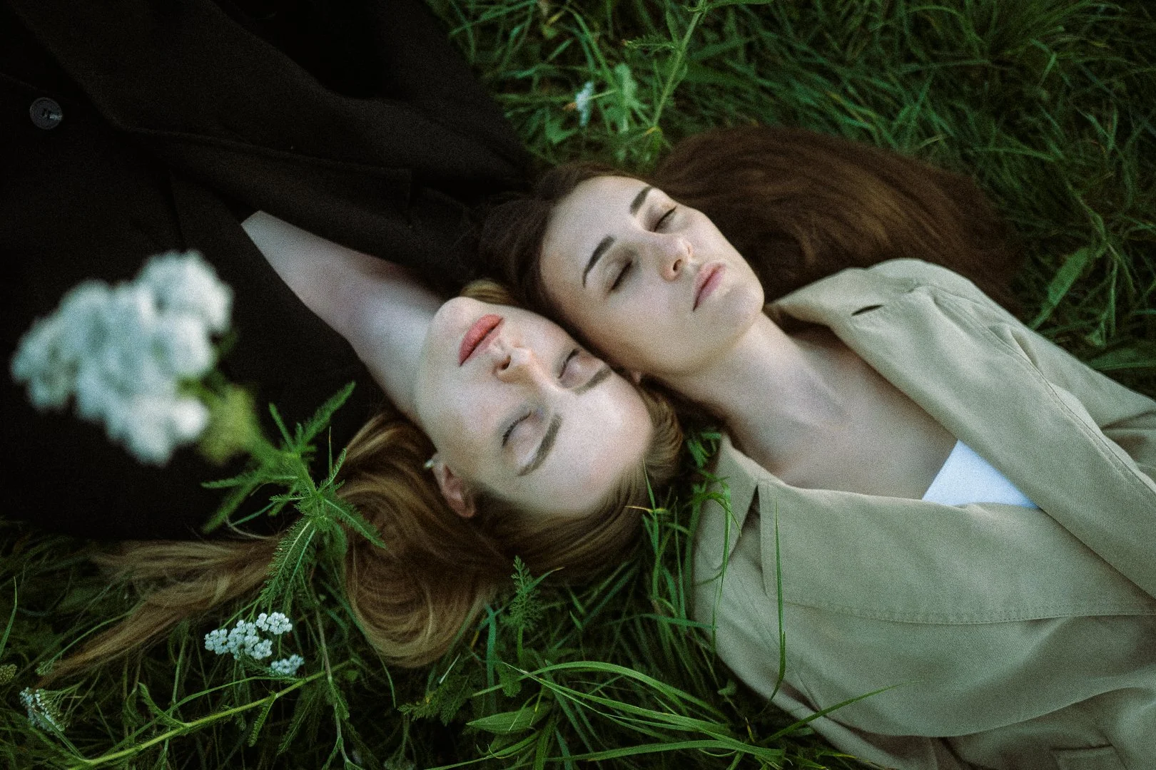 Zwei Frauen liegen mit geschlossenen Augen im Grase, umgeben von grünen Pflanzen, in einer entspannten Pose. Paarshooting der Braut mit ihrer Trauzeugin