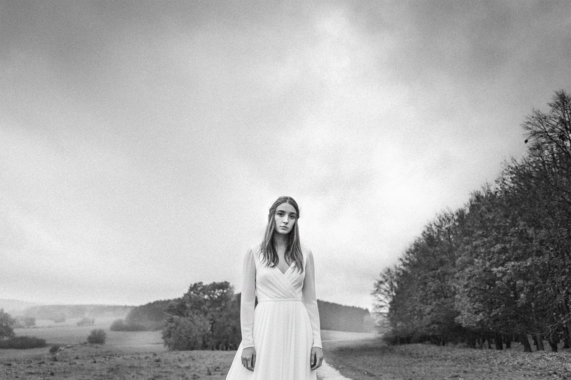 Schwarz-weiß-Foto eines Mädchens in einem weißen Kleid, das auf einer ländlichen Straße steht, umgeben von Bäumen und Hügeln im Hintergrund. Ästhetisches Hochzeitsfoto
