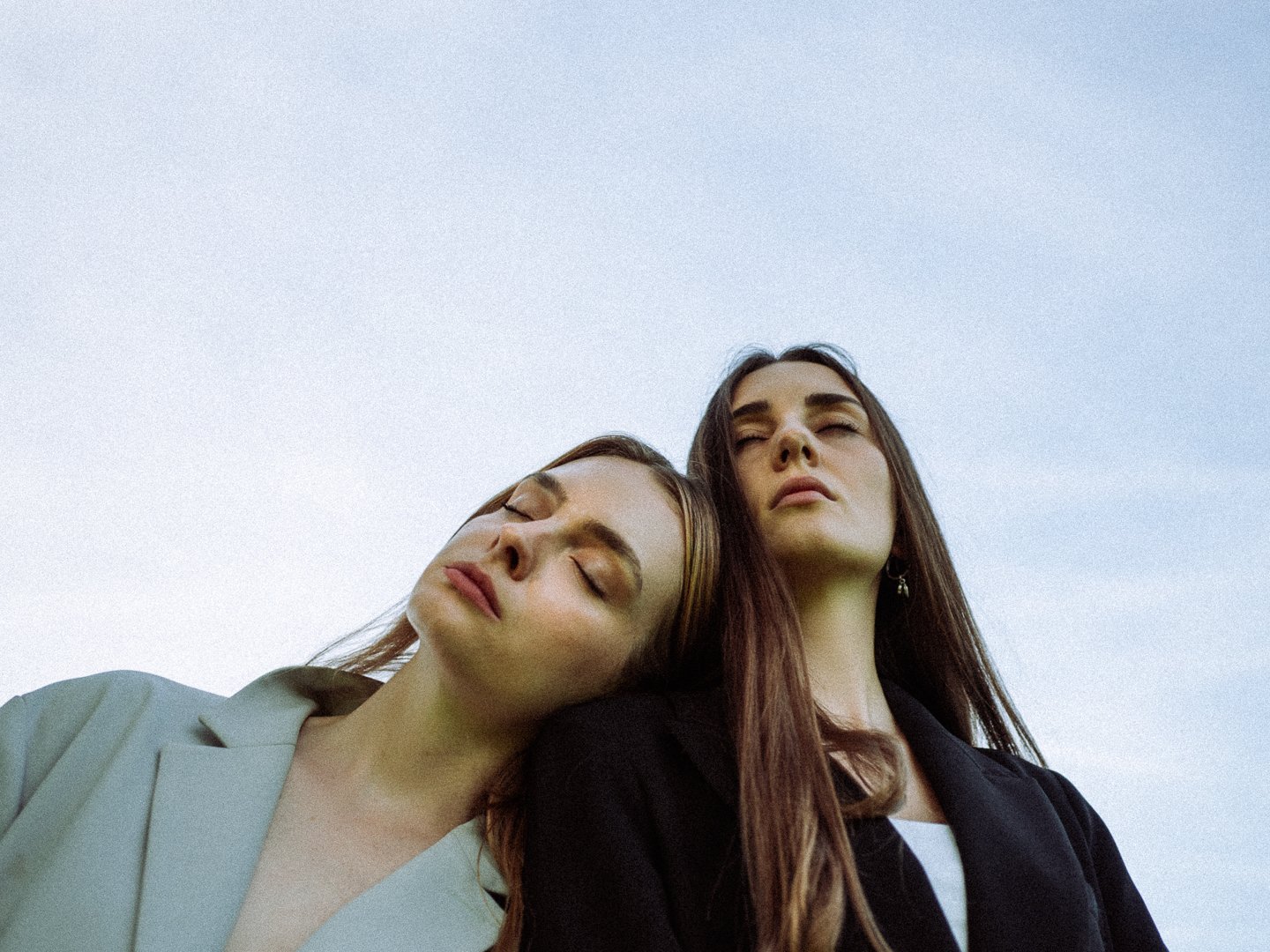 Zwei junge Frauen mit geschlossenen Augen, die ihren Kopf aneinander lehnen, vor einem hellblauen Himmel. Braut und Trauzeugin in vertrauter pose. Emotionale Hochzeitsfotos