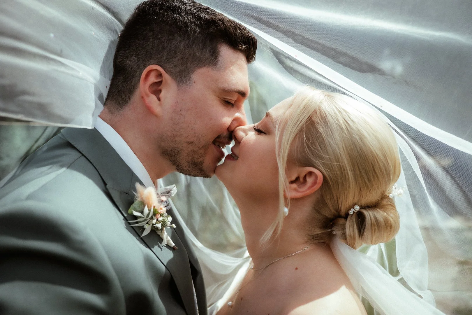 Ein Brautpaar bei ihrer Hochzeitszeremonie, beide stehen sich nahe unter einem weißen Schleier und küssen sich.