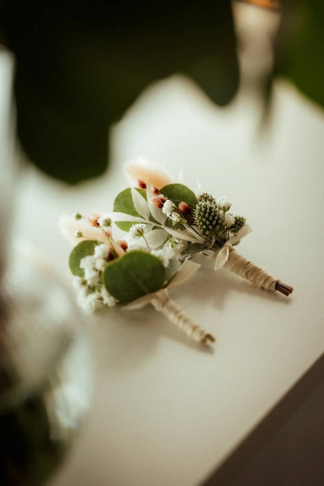 Ein Brautstrauß mit weißen und grünen Blumen liegt auf einer weißen Oberfläche, im Hintergrund sind große grüne Blätter sichtbar.