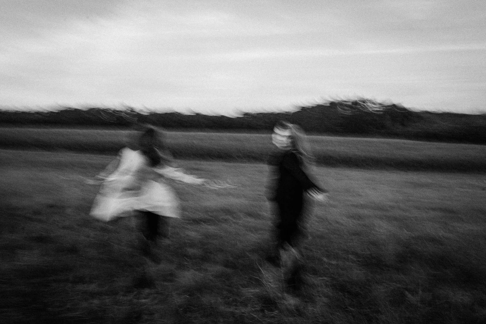 Zwei junge Frauen tanzen im Freien bei Sonnenuntergang auf einem Feld, in Schwarz-Weiß, leicht unscharf, mit ländlicher Landschaft im Hintergrund. Moderne Hochzeitsfotos