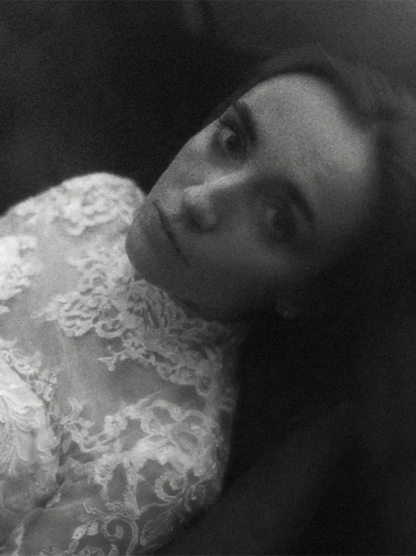 Schwarz-weiße Nahaufnahme einer Frau mit dunklen Haaren, die ein elegantes, lace-besticktes Kleid trägt, in einem dunklen Hintergrund. Emotionale Hochzeitsfotos