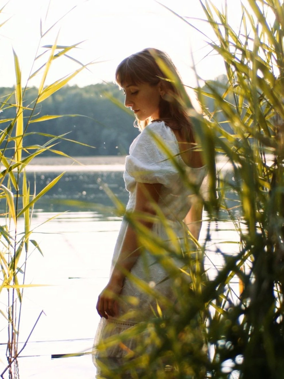 Eine junge Frau im weißen Kleid steht an einem Wasserufer, umgeben von grünen Schilfrohrpflanzen, im Sonnenlicht bei Sonnenuntergang. Intime Hochzeitsfotos