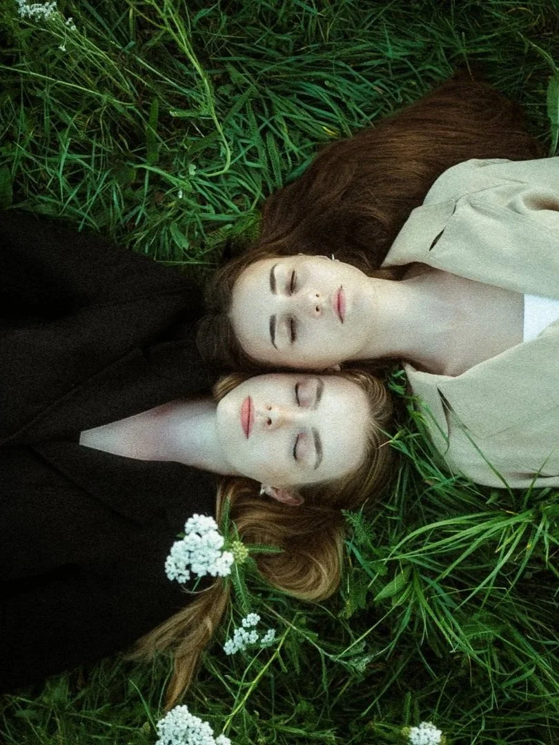 Zwei Frauen mit geschlossenen Augen liegen in hohem Gras, eine trägt dunkle Kleidung, die andere helle Kleidung, mit weißen Blumen im Haar. Emotionale Hochzeitsfotos europa
