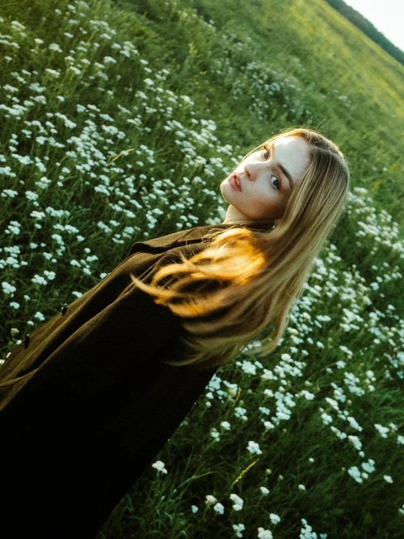 Junge Frau mit langen blonden Haaren steht in einem grünen Feld mit weißen Blumen, Blick zum Himmel, Sonnenlicht auf ihrem Haar, natürliche Umgebung, entspannt. Hochzeitsfotos Bayern