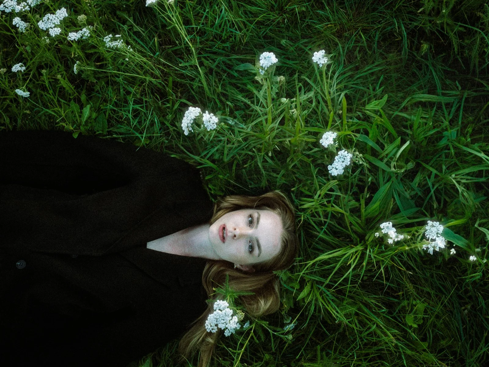 Junge Frau mit langem, leicht gewelltem braunem Haar liegt im Gras mit weißen kleinen Blumen, trägt dunklen Mantel, Blick nach oben, ernster Ausdruck. Moderne Hochzeitsfotos