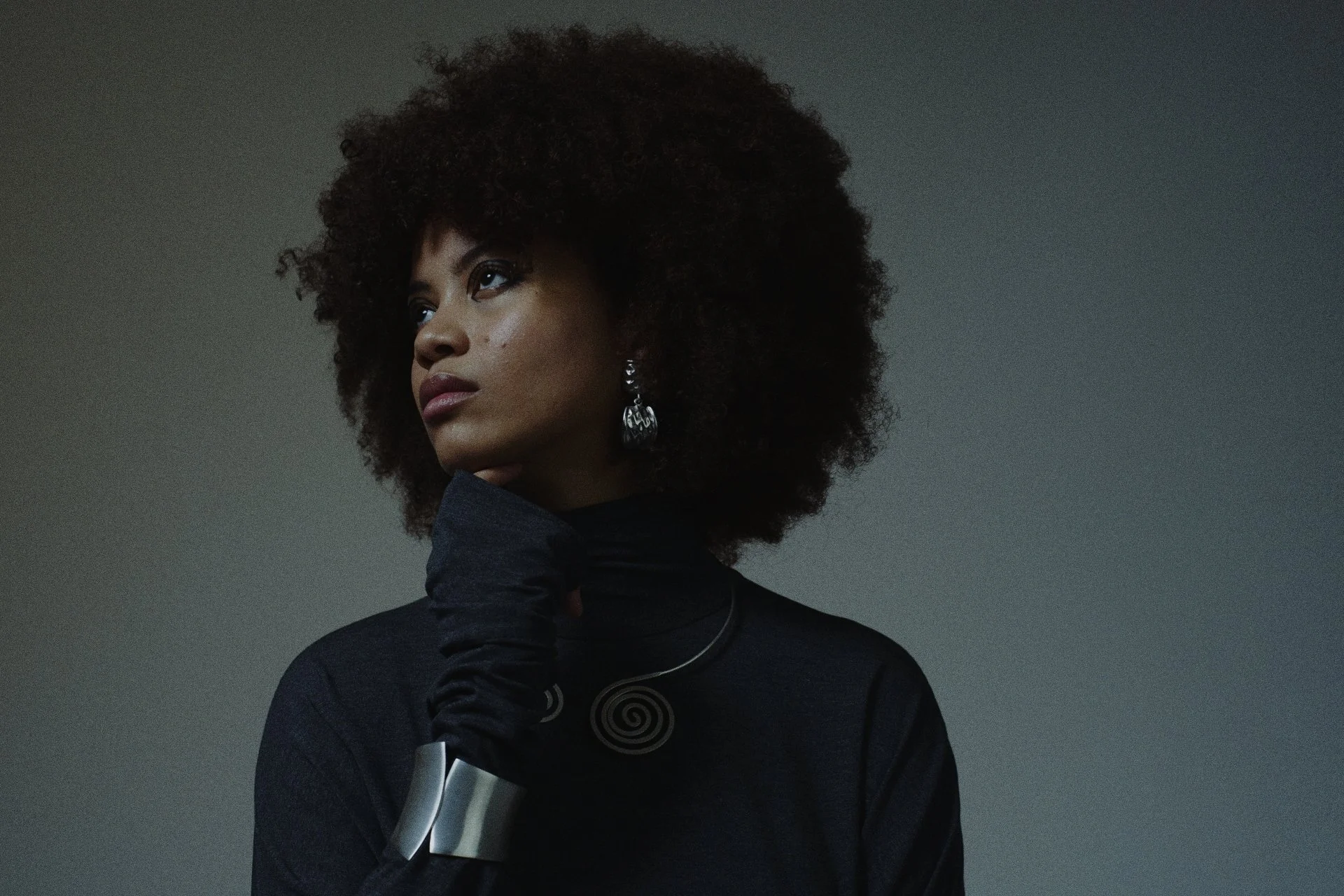 Porträt einer schwarzen Frau mit Afro-Haar, die nachdenklich zur Seite schaut, trägt schwarze Kleidung, silberne Ohrringe, eine Spirale Halskette und ein silbernes Armband. Ästhetische Hochzeitsfotos