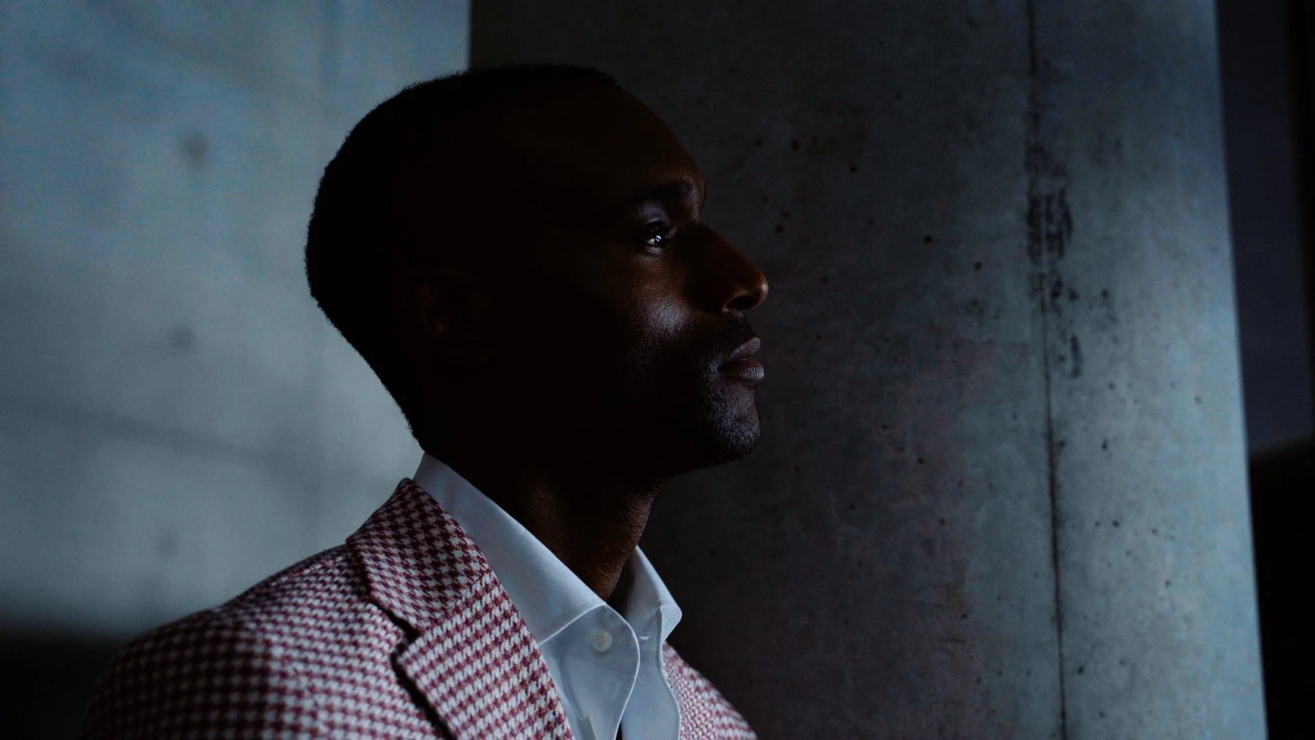 Ein Mann in einem roten und weißen karierten Sakko und weißem Hemd blickt nach rechts, steht vor einer Betonwand, wenig Licht, die Seite des Gesichts sichtbar. Ästhetische Hochzeitsfotos