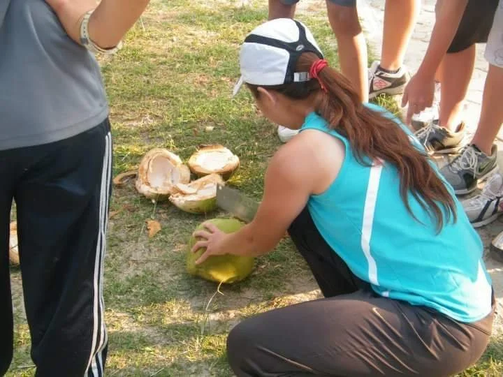 A group of people outdoors, with one woman kneeling down and cutting open a green coconut using a large knife, while others stand around watching.