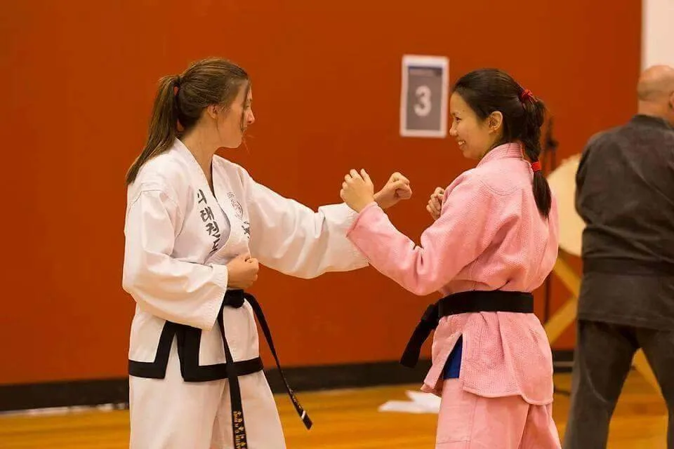 Two women in martial arts uniforms, one in a white gi and the other in a pink gi, practicing martial arts in a dojo. They are holding fists up in a fighting stance and smiling at each other.