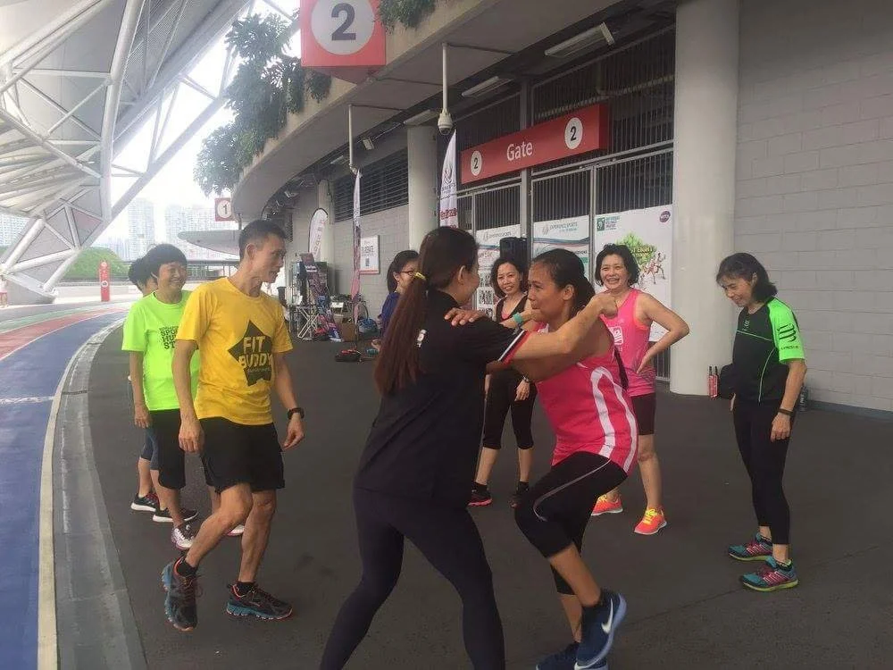Group of women participating in a warm-up or dance session outside a stadium or sports complex near a gate labeled '2'.