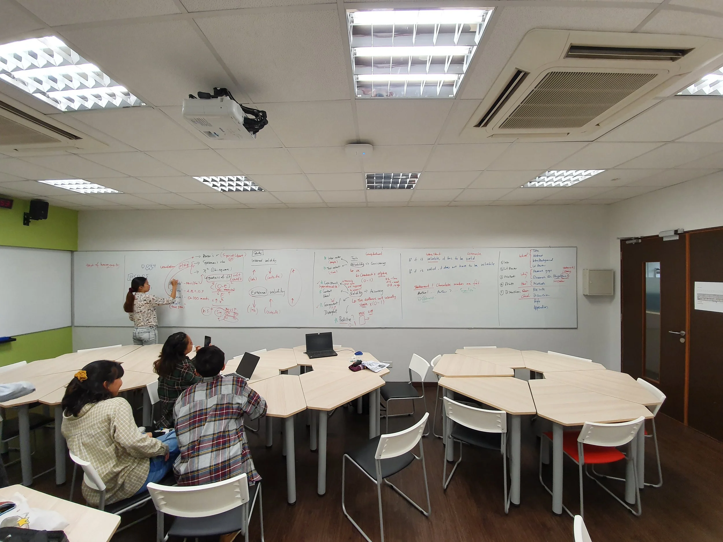 A classroom with a teacher at the whiteboard teaching students seated at tables, some using laptops, in a well-lit room with ceiling lights and air conditioning.