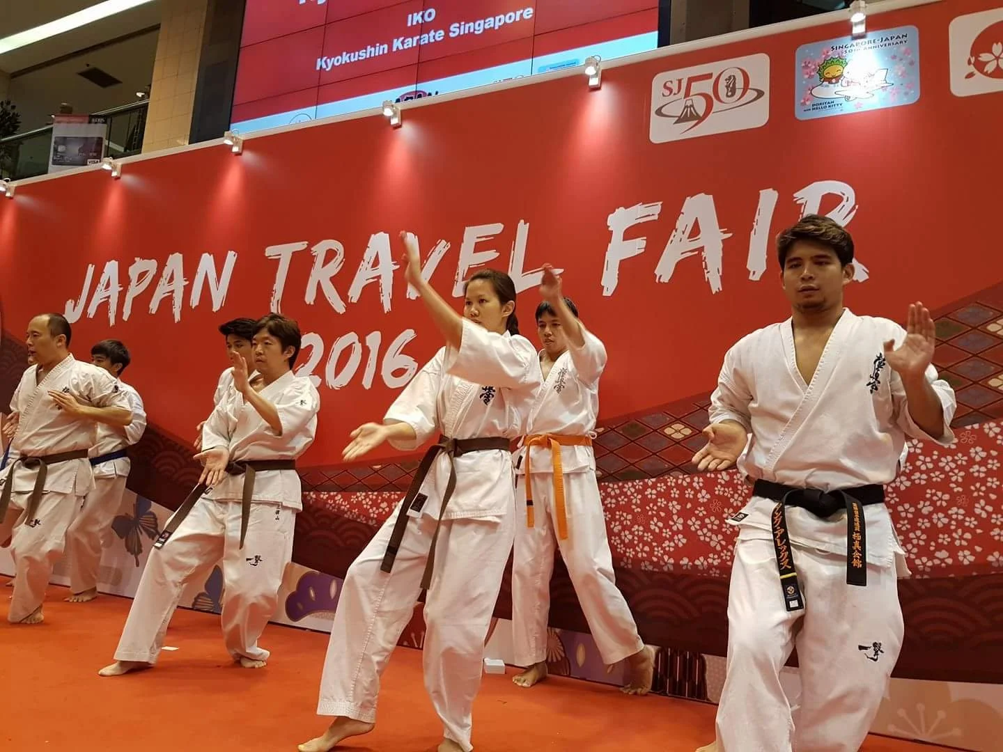 Group of martial artists practicing karate on stage at Japan Travel Fair 2016, wearing traditional white gis with black belts, in front of a large red backdrop with the event name and logos.